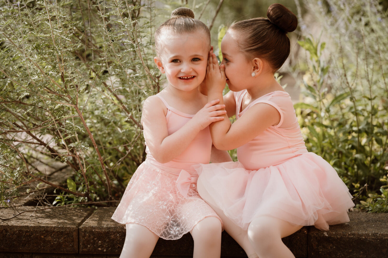 Two little girls in pink dresses are sitting on a bench