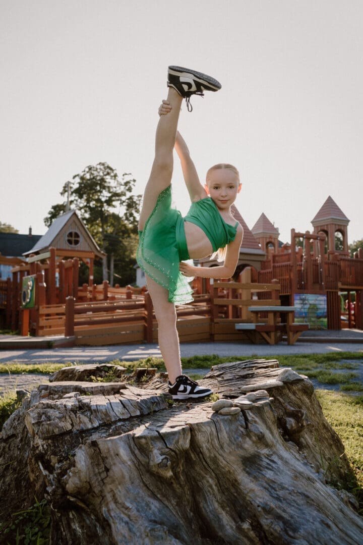 A young girl in a green tutu performs a high leg stretch on a tree stump.