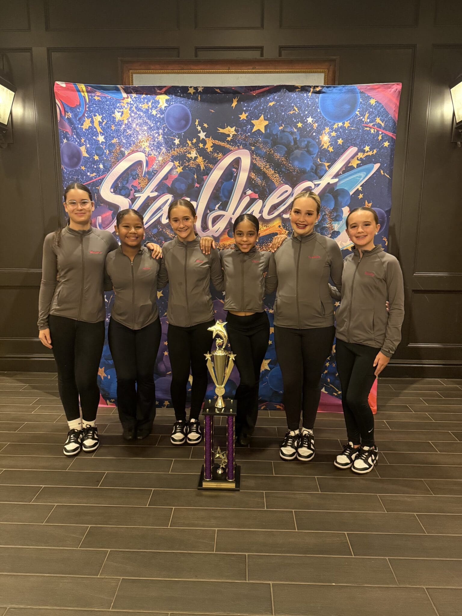 A group of seven young dancers posing with a large trophy indoors.